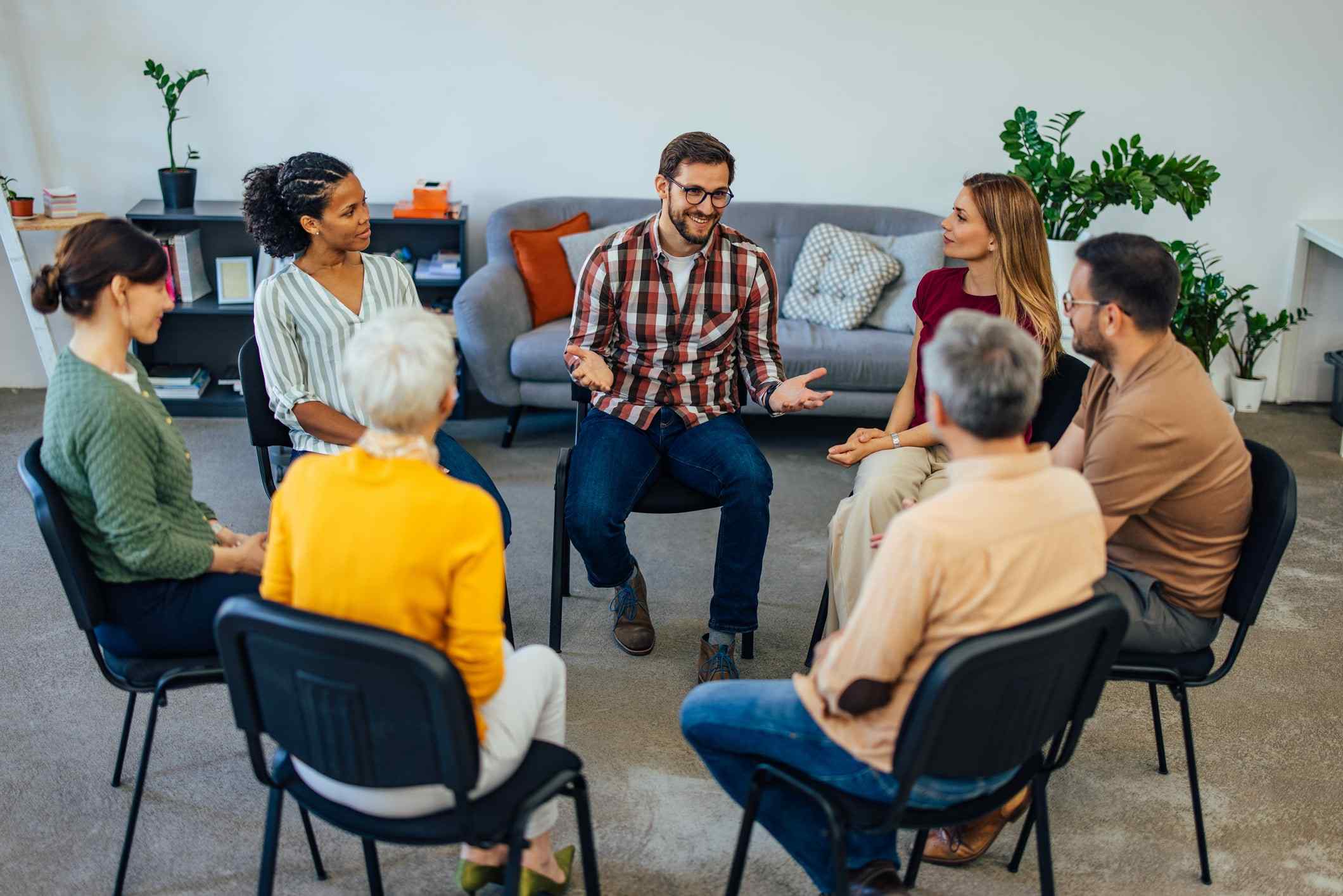 A diverse group of people sit in a circle during a casual discussion. They appear engaged and relaxed, with a sofa and plants in the background.