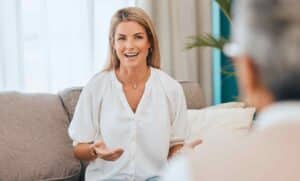 A smiling woman in a white blouse sits on a sofa, gesturing with her hands during a conversation. The atmosphere is relaxed and engaging.