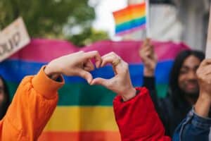 Hands forming a heart against a vibrant rainbow flag backdrop, surrounded by people holding rainbow flags. The scene conveys unity and pride.