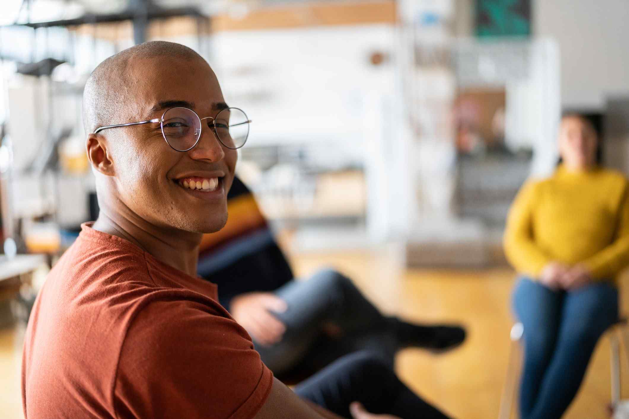 A person with glasses smiles warmly in a relaxed, casual group setting. The atmosphere is friendly, with soft lighting and blurred background figures.