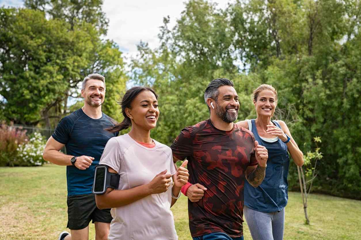 Group of four adults jogging in a park, smiling and wearing athletic gear. They appear happy and energetic, surrounded by green trees under a clear sky.