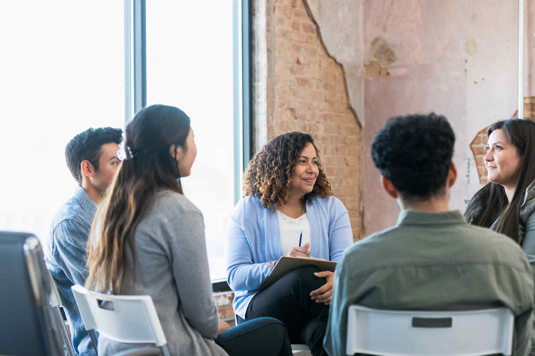 A diverse group of five people sit in a circle, engaged in a positive discussion. The woman in the center smiles, holding a clipboard, inside a rustic room.