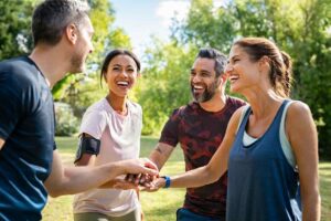 Four people in sportswear stand in a circle outdoors, laughing and shaking hands in a sunny park. The scene is cheerful and friendly.