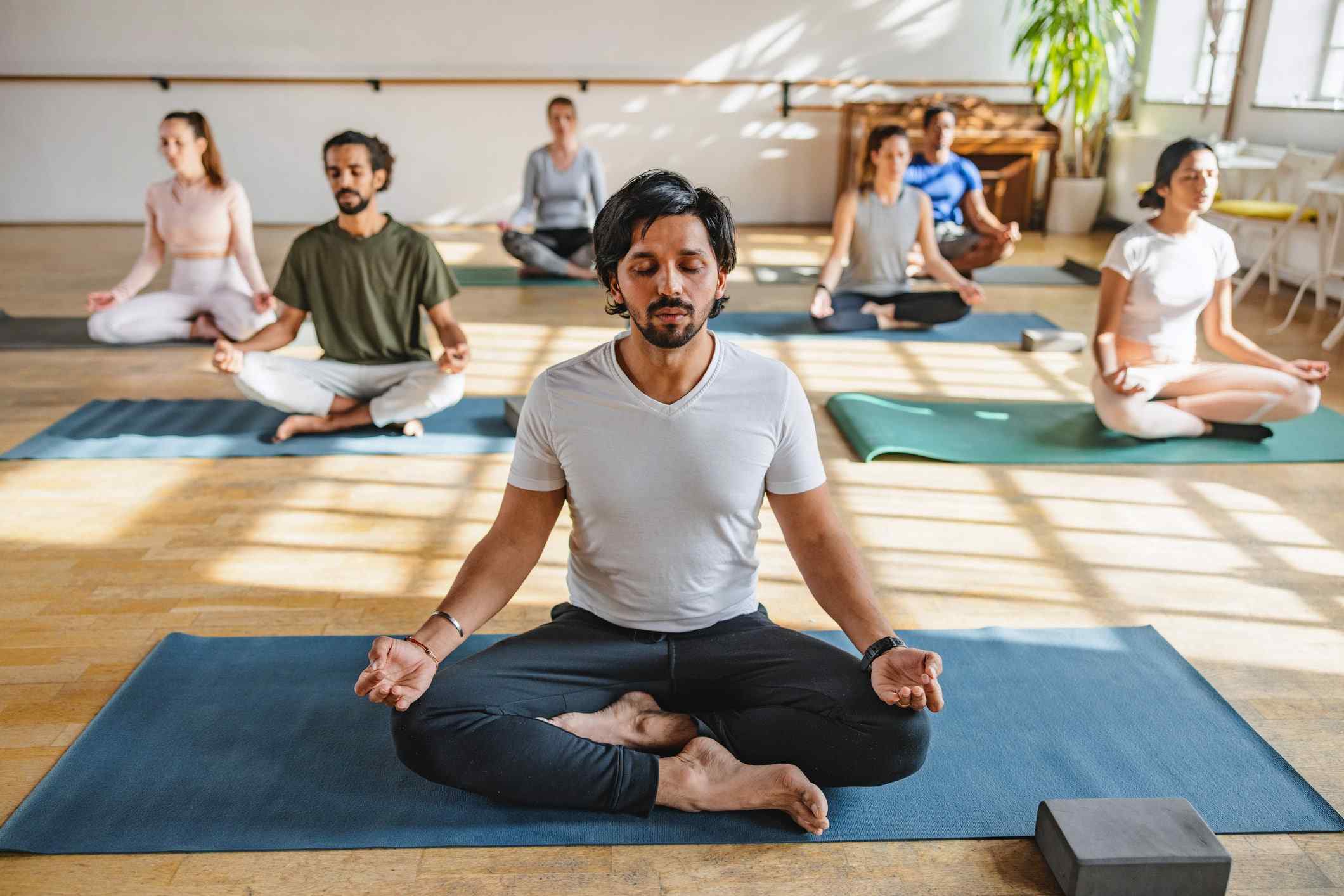 Group of men and women practicing meditation in lotus pose on yoga mats inside a bright studio with wooden floors and large windows, led by a man in a white t-shirt and black pants seated at the front, all with eyes closed and hands resting on knees in a mindful posture. Ask ChatGPT