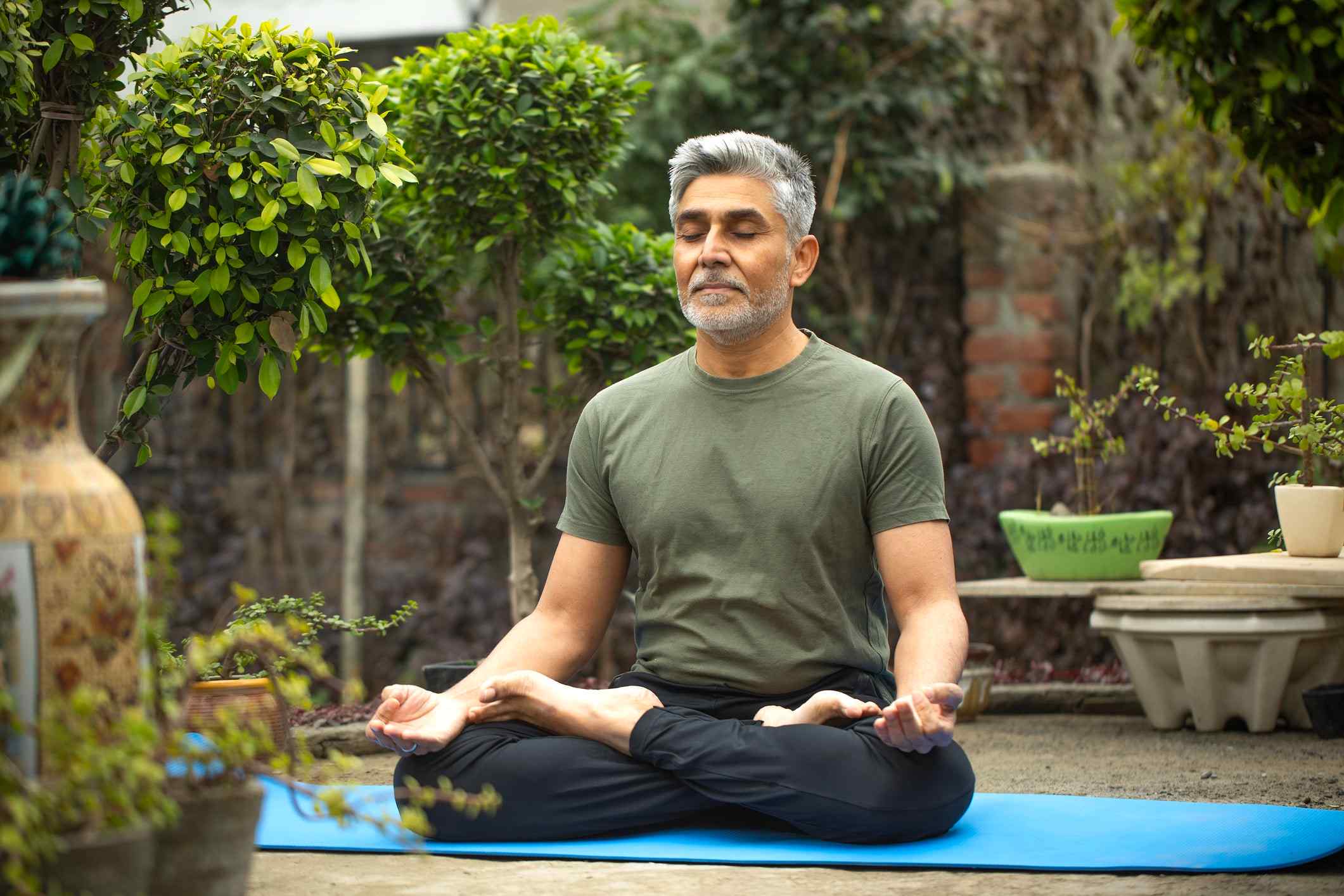 Middle-aged man with short gray hair and beard practicing meditation in lotus pose on a blue yoga mat in an outdoor garden, wearing a green t-shirt and black pants, surrounded by lush green plants, potted shrubs, and decorative vases, eyes closed in peaceful mindfulness practice. Ask ChatGPT