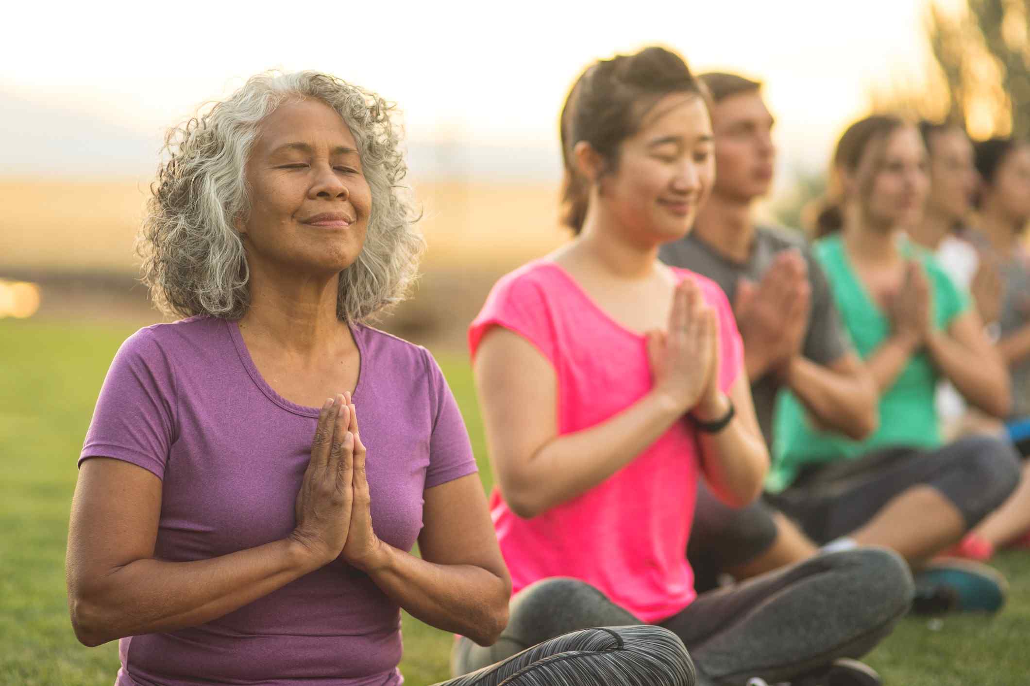 A group of people practicing yoga outdoors, seated with hands in a prayer position, surrounded by lush greenery and a sunset backdrop.