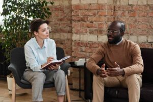 A woman in a light blue shirt attentively takes notes while listening to a man in a brown sweater speaking. They are sitting in a cozy room with a brick wall backdrop, conveying a professional yet relaxed atmosphere.