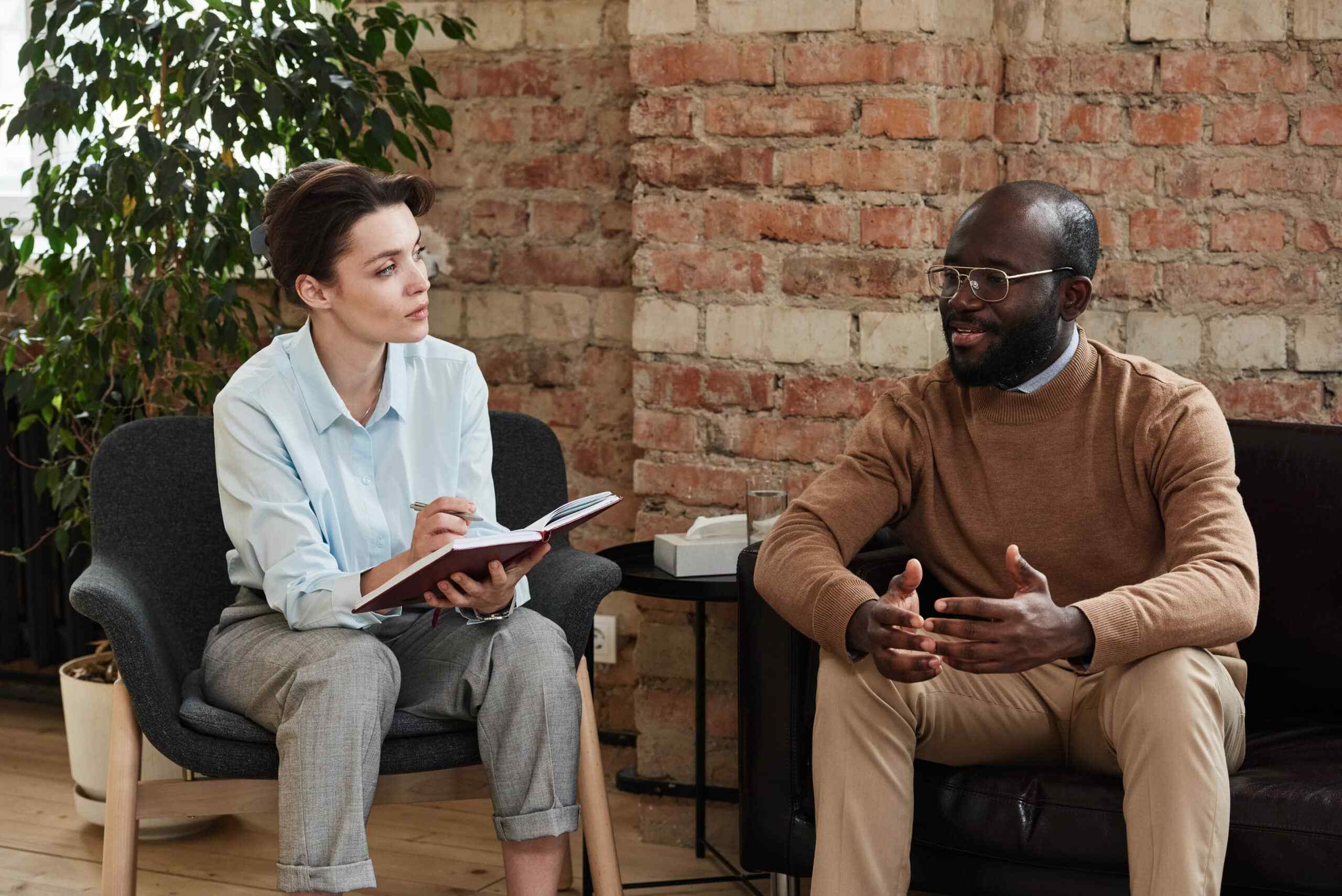 A woman in a light blue shirt attentively takes notes while listening to a man in a brown sweater speaking. They are sitting in a cozy room with a brick wall backdrop, conveying a professional yet relaxed atmosphere.