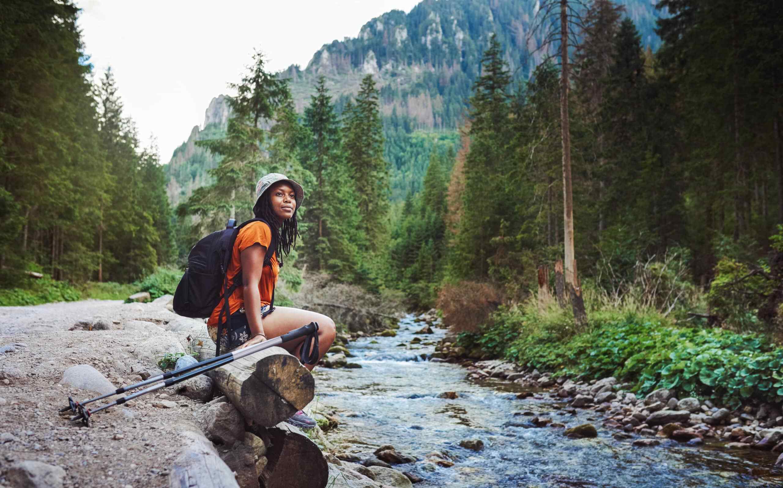 A woman in hiking attire sits on a log beside a flowing stream, surrounded by tall, dense pine trees and mountains, conveying a serene and adventurous tone.