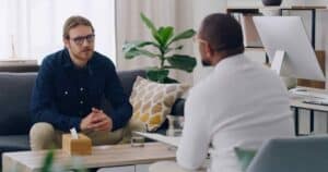 Two men sit in a cozy office, engaged in a serious conversation. One man listens intently, while the other speaks. There's a plant and a computer in the background.