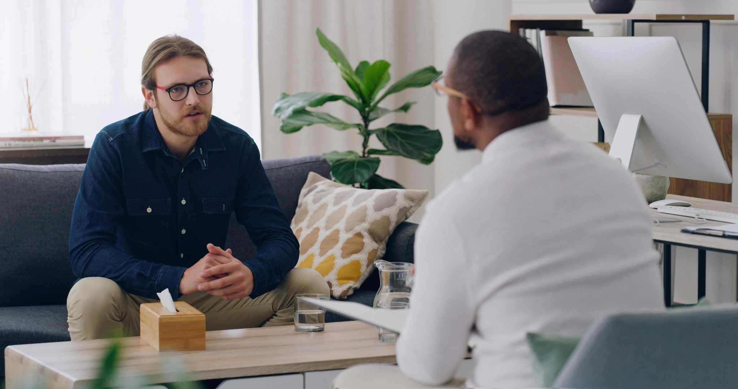 Two men sit in a cozy office, engaged in a serious conversation. One man listens intently, while the other speaks. There's a plant and a computer in the background.