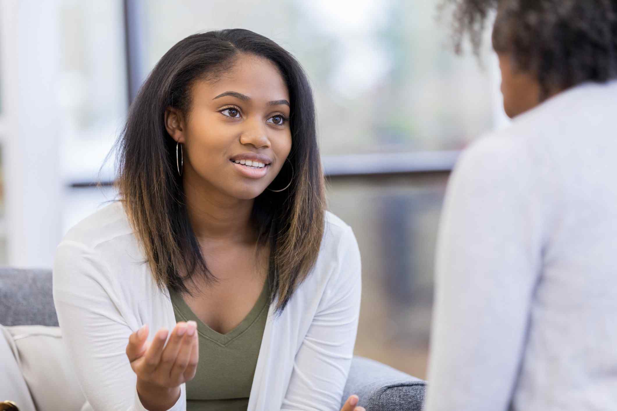 A woman with long hair, wearing a white cardigan, attentively listens and gestures while sitting on a couch, engaging in a conversation with another person.