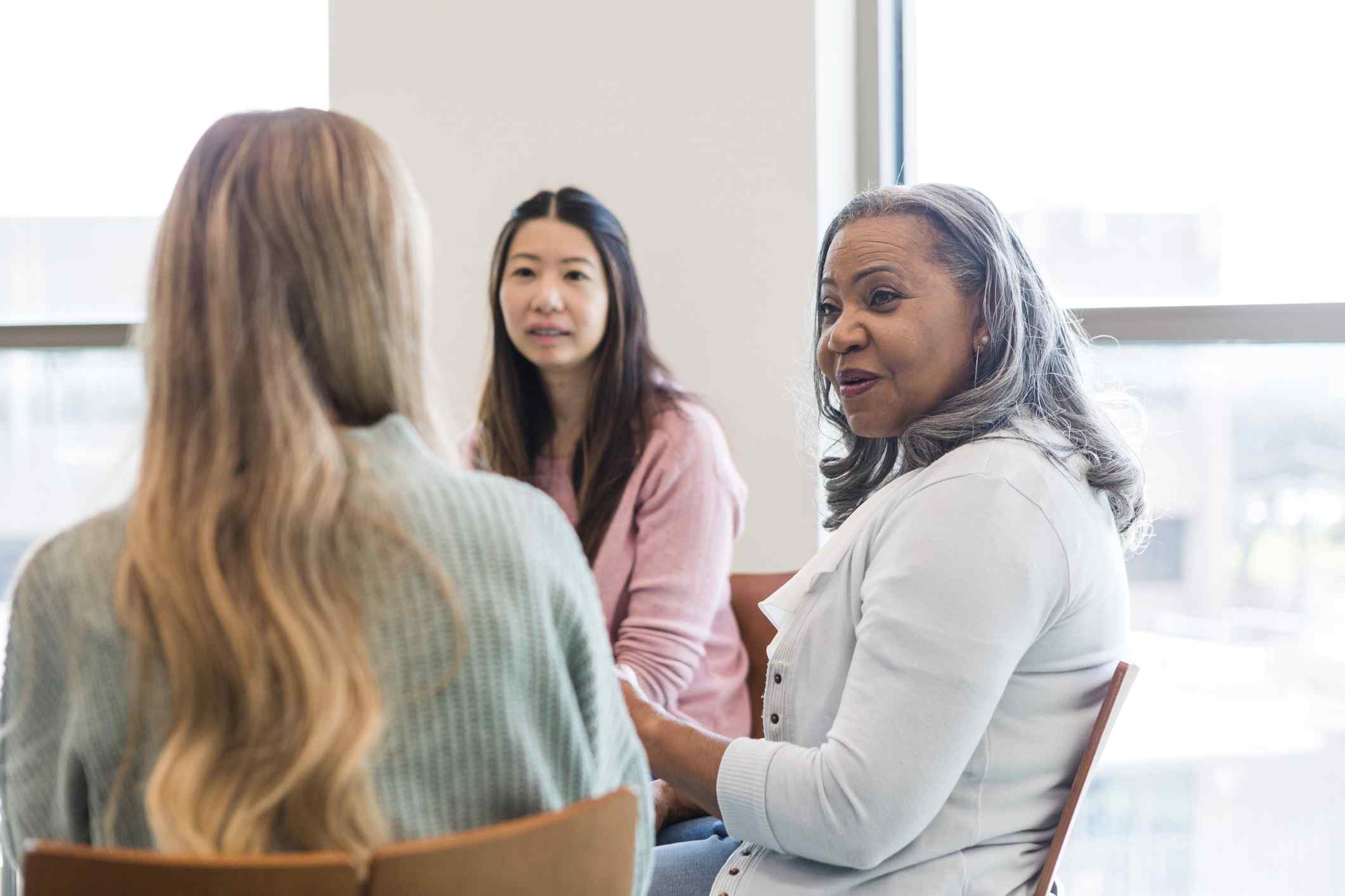 Three women sit in a casual meeting, engaged in conversation near a window. The atmosphere is friendly and collaborative, with natural lighting.