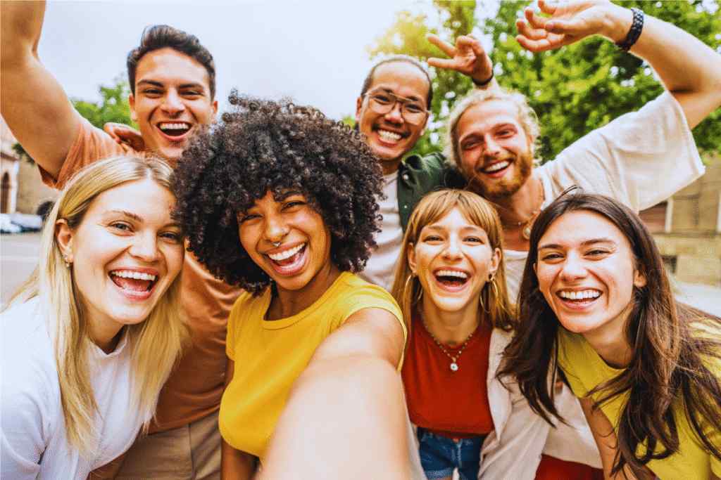 A group of cheerful friends posing for a selfie outdoors, surrounded by greenery, wearing casual colorful clothing and smiling.