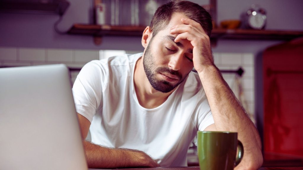 The What & Why Behind Alcoholism: Alcohol Use Disorder Explained 2 Person holding their head at a kitchen table, showing physical and mental strain associated with alcohol withdrawal.